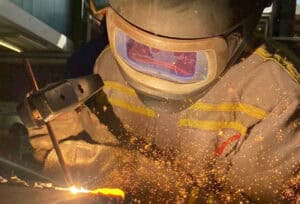 A metal welder at work, cutting and welding in an industrial setting, wearing safety goggles and work clothes.
