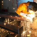 Ein Schweißer arbeitet in einer Werkstatt, verschweißt Metallteile und erzeugt Funken, während er eine Schutzbrille und Arbeitskleidung trägt.